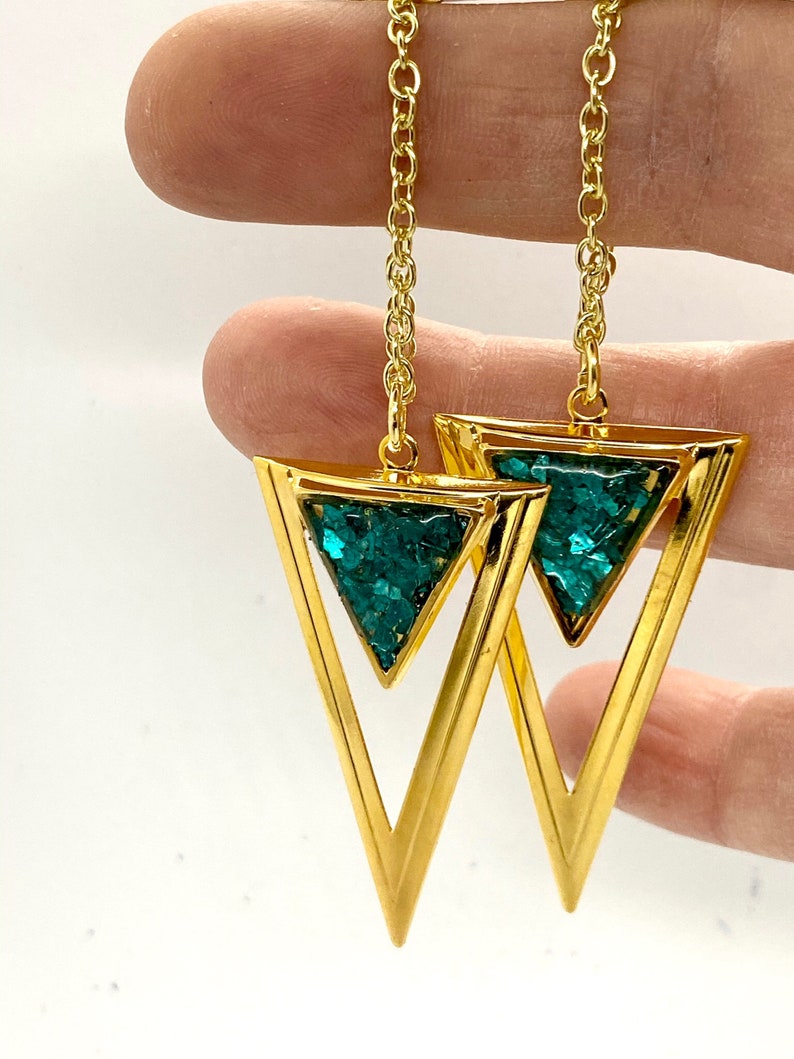May include: Gold-tone earrings featuring open triangle pendants filled with sparkling teal-colored material. The earrings hang from delicate gold chains. The design is geometric and modern, suitable for everyday wear or special occasions.
