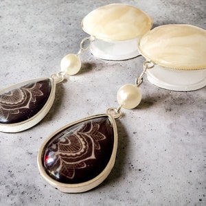 May include: A pair of dangle earrings with a silver-tone setting. The teardrop-shaped pendants feature a dark burgundy background with a white floral design. Each earring has a white pearl and a white disc-shaped top.