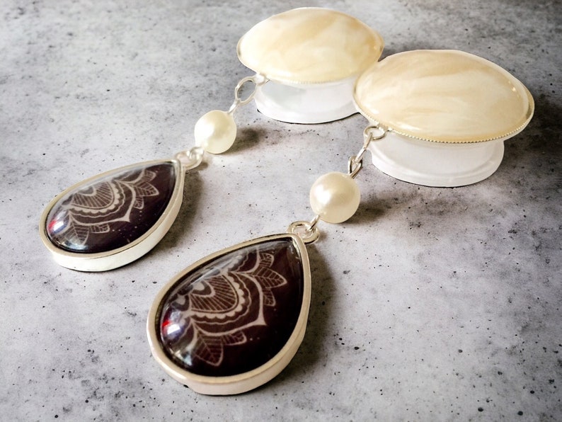 May include: A pair of dangle earrings with a silver-tone setting. The teardrop-shaped pendants feature a dark burgundy background with a white floral design. Each earring has a white pearl and a white disc-shaped top.