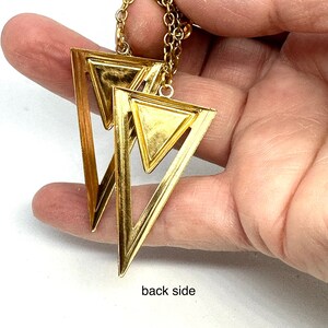 May include: Gold-toned geometric earrings featuring a layered triangle design. The earrings are suspended from a delicate chain. The image shows the back side of the earrings. The earrings are held in a hand.