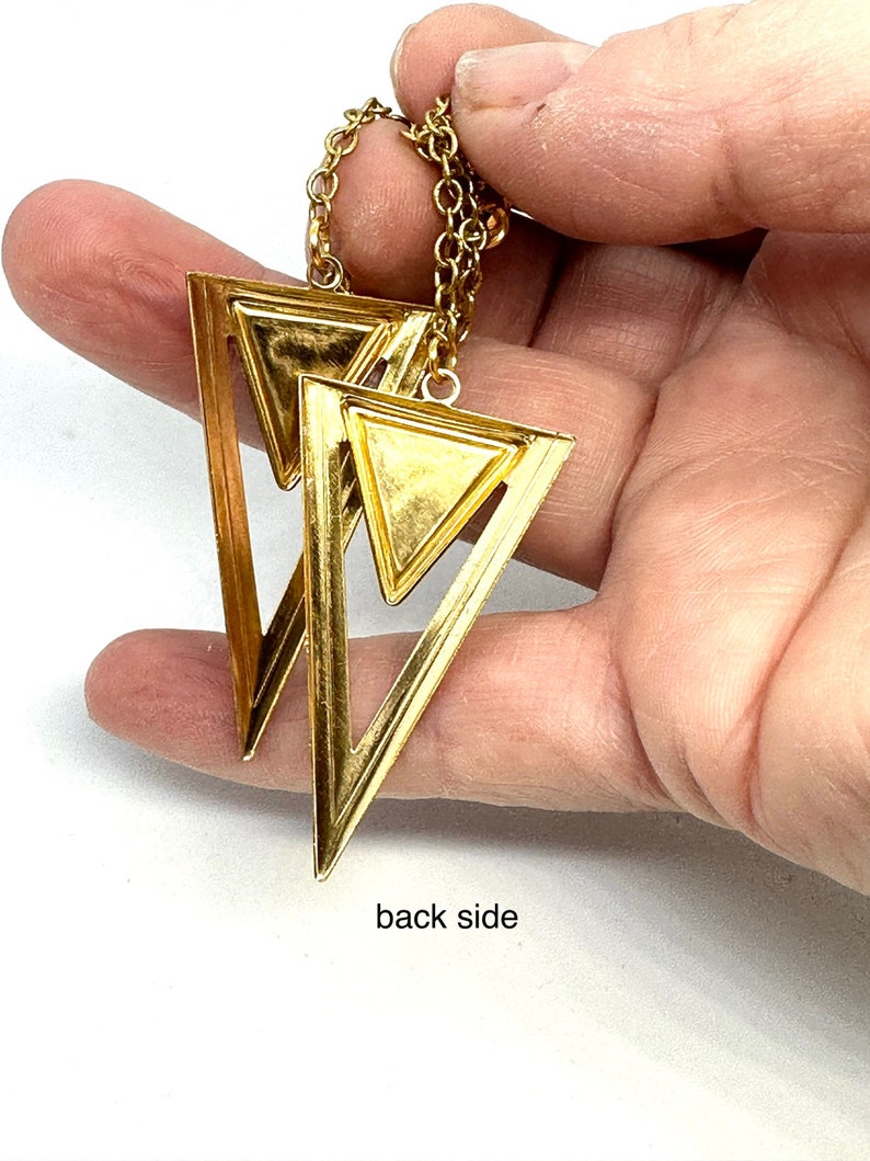 May include: Gold-toned geometric earrings featuring a layered triangle design. The earrings are suspended from a delicate chain. The image shows the back side of the earrings. The earrings are held in a hand.