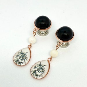 Puede incluir: Un par de pendientes colgantes de rosa negra y blanca con una piedra negra y una perla blanca. Los pendientes están hechos de acero inoxidable y tienen un acabado en oro rosa.