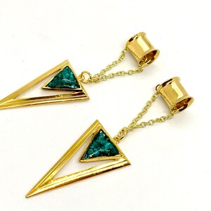 May include: A pair of gold-colored earrings featuring a geometric design. Each earring has a triangular shape with a turquoise-colored stone inlay and a chain connecting to a cylindrical post.