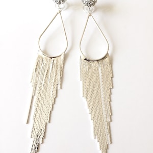 Op de afbeelding: Een paar zilverkleurige oorhangers. Elke oorbel heeft een kleine, ronde, met strass bezette stud, verbonden met een druppelvormig frame. Onder het frame hangt een waterval van zilveren franjes, wat een glamoureus, opvallend ontwerp creëert.