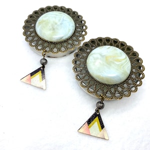 May include: A pair of white and gold ear gauges with a decorative filigree design and a dangling triangle charm. The gauges feature a white marble-like pattern.