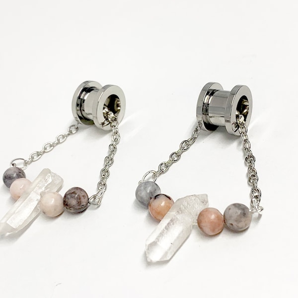Op de afbeelding: Een paar zilveren roestvrijstalen oorpluggen met een ketting en kristal- en roze en grijze kralen die eraan hangen.