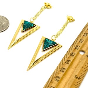 May include: A pair of gold-colored earrings with a geometric design. Each earring features a gold triangle with a turquoise-colored stone inlay. The earrings hang from a delicate gold chain. A ruler shows the earrings are approximately 2.5 inches long.