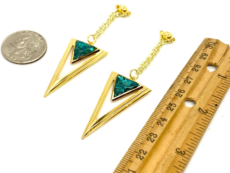 May include: A pair of gold-colored earrings with a geometric design. Each earring features a gold triangle with a turquoise-colored stone inlay. The earrings hang from a delicate gold chain. A ruler shows the earrings are approximately 2.5 inches long.