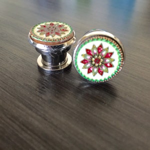 May include: Two decorative cabinet knobs with a floral design. The knobs have a silver base and a round top with a white background and a red, green, and gold floral pattern. The knobs are on a dark wood surface.