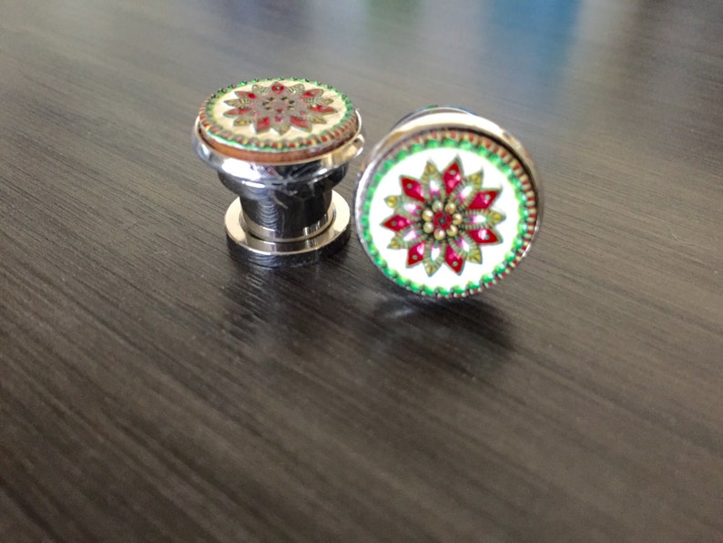 May include: Two decorative cabinet knobs with a floral design. The knobs have a silver base and a round top with a white background and a red, green, and gold floral pattern. The knobs are on a dark wood surface.