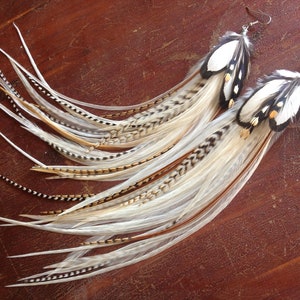 Puede incluir: Un par de pendientes largos de plumas blancas, marrones y negras. Los pendientes están hechos con ganchos de plata.