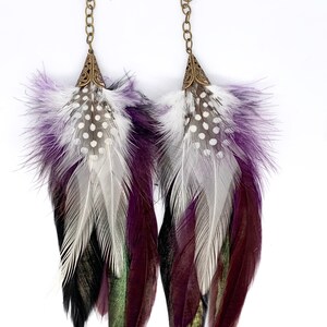 Puede incluir: Un par de pendientes largos de plumas con un esquema de color morado, blanco, negro y verde. Los pendientes tienen una cadena de latón y una parte superior de latón decorativa.