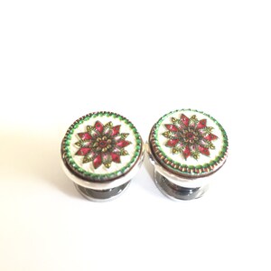 May include: Two decorative ear plugs with a floral design. The plugs have a white background with a red and gold floral pattern, a green border, and a silver base. The plugs are round and symmetrical.