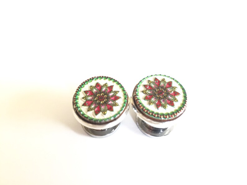 May include: Two decorative ear plugs with a floral design. The plugs have a white background with a red and gold floral pattern, a green border, and a silver base. The plugs are round and symmetrical.