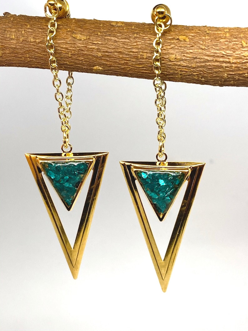 May include: A pair of gold-colored earrings with a geometric design. Each earring features a gold triangle frame with a smaller triangle filled with teal-colored glitter. The earrings hang from a gold chain.