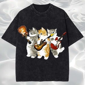Puede incluir: Camiseta negra con un gráfico de tres gatos de dibujos animados tocando la guitarra. Los gatos son grises, naranjas y grises con detalles blancos. Cada gato sostiene una guitarra de diferente color. La camiseta tiene un aspecto vintage y lavado.