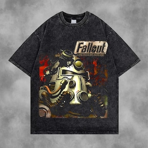 May include: Dark gray t-shirt featuring a distressed graphic of a power armor suit from the Fallout video game series. The shirt has the word "Fallout" printed above the graphic. The shirt is made of a soft, comfortable material.