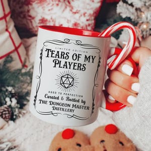 Peut inclure: Mug en céramique blanche avec intérieur et anse rouges, avec le texte "TEARS OF MY PLAYERS" et un graphisme sur le thème de D&D. Un sucre d'orge rouge et blanc est dans la tasse. La tasse est de "THE DUNGEON MASTER DISTILLERY".