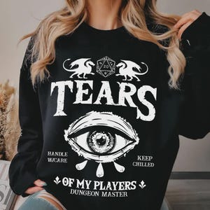 Può includere: Felpa nera con un design grafico bianco. Il design include la parola "TEARS" sopra un'illustrazione dettagliata di un occhio. Sotto l'occhio, si legge "OF MY PLAYERS DUNGEON MASTER". Due draghi e un dado D20 sono sopra la parola "TEARS".