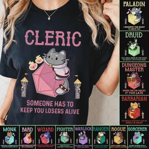 Puede incluir: Camiseta negra con un diseño rosa "Cleric" y un gato de dibujos animados sosteniendo una gema rosa. El texto "Someone has to keep you losers alive" está debajo. Gráficos adicionales representan varias clases de D&D.