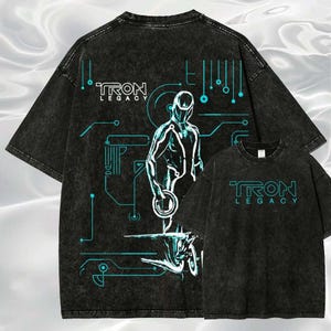 May include: Black acid wash t-shirts with a Tron Legacy design. The front and back of the shirts feature a white and teal graphic of a figure and circuit board elements. The text "TRON LEGACY" is also displayed.