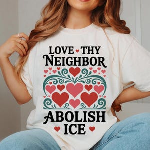 May include: A cream-coloured t-shirt featuring the text "LOVE THY NEIGHBOR ABOLISH ICE" in black. The design incorporates red and pink hearts, and green decorative elements. This is a casual wear item.