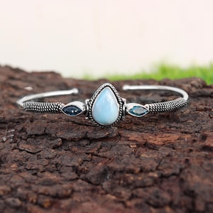 May include: A silver-toned cuff bracelet featuring a pear-shaped, light blue gemstone at the center, framed by a twisted rope design. Two smaller blue gemstones flank the center stone. The bracelet rests on a textured, brown surface.