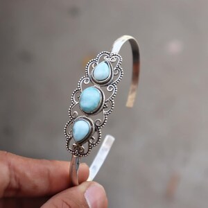 May include: Silver cuff bracelet featuring three light blue teardrop-shaped gemstones. The gemstones are set in ornate, filigree-style settings. The bracelet has an open design and is held by a hand.