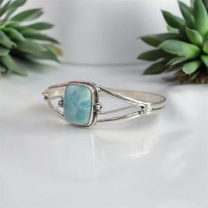 May include: A silver bracelet featuring a rectangular, light blue gemstone. The bracelet has a delicate, open design with curved lines and small decorative elements. The gemstone is the focal point, set in a silver bezel. The bracelet is displayed on a white surface.