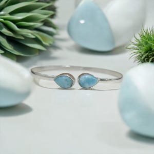 May include: A silver-toned cuff bracelet featuring two pear-shaped, light blue gemstones. The bracelet is set against a white surface with decorative elements in the background, including a succulent plant and light blue spheres.