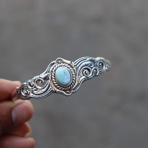 May include: Silver-toned bracelet featuring an oval, light blue gemstone at its center. The bracelet has an ornate, swirling design around the gemstone and along the band. The jewelry is displayed against a blurred background.