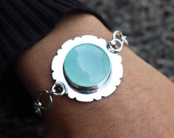May include: A silver bracelet with a large, round, pale blue gemstone set in a scalloped bezel. The bracelet has a delicate chain with circular links. The gemstone is the focal point, with a polished, smooth surface.