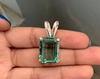May include: A rectangular, teal-colored gemstone pendant with a silver bail. The gemstone is set in a silver frame and has a clear, transparent appearance. The pendant is held in a hand.