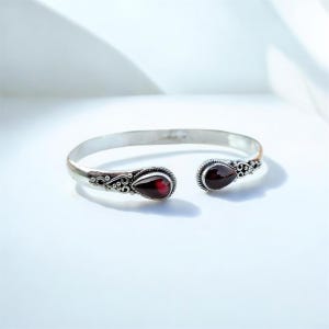 Puede incluir: Brazalete de plata con detalles ornamentados y dos gemas rojas oscuras en forma de lágrima. El brazalete de diseño abierto se exhibe sobre una superficie blanca, mostrando la elegante artesanía de la joya.