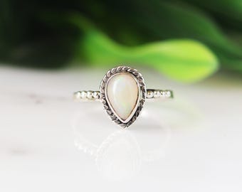 Könnte beinhalten: Ein silberner Ring mit einem birnenförmigen, cremefarbenen Opal-Edelstein. Der Edelstein ist in einer dekorativen Lünette mit einem seilartigen Design gefasst. Das Band hat kleine, perlenartige Akzente. Der Ring wird auf einer weißen Oberfläche mit verschwommenem grünem Hintergrund gezeigt.