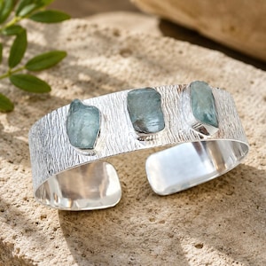 Peut inclure: Un bracelet manchette en argent présentant une finition texturée semblable à de l'écorce sur la surface extérieure. Le bracelet est orné de trois pierres brutes bleu-vert clair serties dans des chatons en argent individuels.