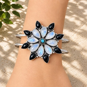 Può includere: Un bracciale a fascia in argento con un grande elemento centrale a forma di fiore. Il design presenta pietre di luna e onice nero a goccia disposte a raggiera, con una singola pietra rotonda blu iridescente posizionata al centro.