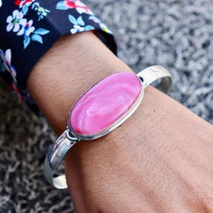 Puede incluir: Una pulsera de plata con una gran piedra rosa ovalada. La piedra tiene un aspecto veteado. La pulsera se lleva en la muñeca, con un fondo gris borroso. El brazalete es un diseño sencillo y elegante.