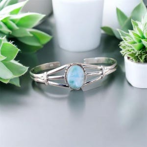 May include: A silver cuff bracelet with an oval, light blue gemstone center. The bracelet has a simple, open design with three parallel bars on each side of the gemstone. The gemstone has a marbled appearance with shades of blue and white.