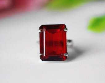 Könnte beinhalten: Ein silberner Ring mit einem großen, rechteckigen, tiefroten Edelstein. Der Edelstein wird von vier Krappen gehalten, und der Ring ist aus schlichtem Silber. Der Hintergrund ist verschwommen, mit Hauch von Rosa und Grün.