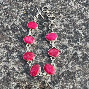 Puede incluir: Una pulsera de plata con siete piedras preciosas rosas ovaladas. Las piedras están engastadas en biseles individuales de plata y conectadas por eslabones de plata. La pulsera tiene un cierre de palanca. La joya se exhibe sobre una superficie texturizada.