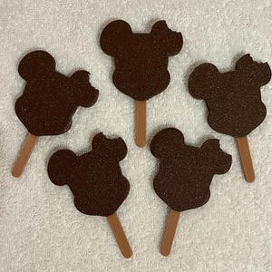 May include: Five dark brown Mickey Mouse-shaped ice cream treats on wooden sticks, some with bite marks, arranged on a white textured surface. The treats have a glittery texture, suggesting a chocolate coating.