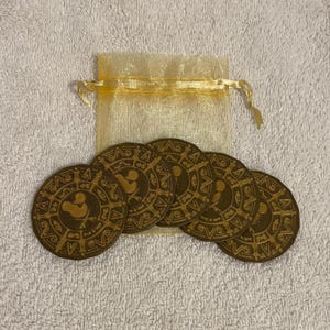 May include: A collection of six brown, circular coins with a decorative design and a cartoon character emblem, next to a small, sheer, gold drawstring bag. The coins are arranged on a white, textured surface.