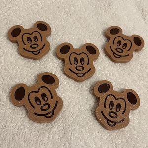 Peut inclure: Cinq objets bruns en forme de Mickey Mouse avec des accents brun foncé. Chaque objet a un visage souriant et de grandes oreilles. Les objets sont disposés sur une surface blanche et texturée.