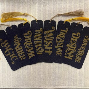 May include: A collection of seven navy blue bookmarks with gold lettering and tassels. Each bookmark features a different word: Magic, Wonder, Dream, Fantasy, Wish, Treasure, Destiny, and Adventure. The tassels are gold and tan.
