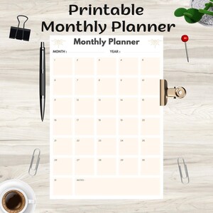 May include: A printable monthly planner with a cream-colored grid, held by a gold clip. The planner has the title "Printable Monthly Planner" at the top. A black pen, paper clips, and a cup of coffee are also visible.