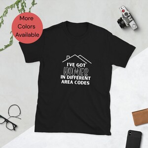 May include: Black t-shirt with the text "I've Got Homes in Different Area Codes" in white. The design includes a house outline above the text. A red circle with the text "More Colors Available" is in the upper left corner.