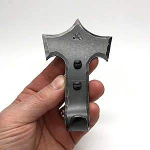 May include: A black metal hook with a unique, star-shaped top. The hook is hand-forged and has a rustic, industrial look. The hook is attached to a wall with two screws.