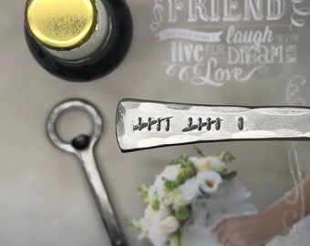 May include: A silver bottle opener with a handle that says "Mr. & Mrs. 1" and a ring on the top. The bottle opener is on a white surface with a wedding card in the background. The card says "Today I married my best friend & together we will laugh, live for dreams & love."