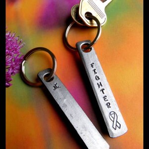 May include: Two silver keychains with key rings. One keychain has the word "Fighter" engraved on it with a ribbon design. The other keychain has the initials "J.C." engraved on it.